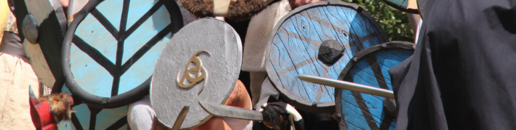 L'image montre un gros plan sur un mur de bouclier dans une scène de combat. On y voit plusieurs blasons sur les boucliers, quatre bleus avec des traits noirs, un gris avec le symbole de la triple corne d'Odin en dorée et un jaune et noir. On peut aussi apercevoir des épées en mousse.