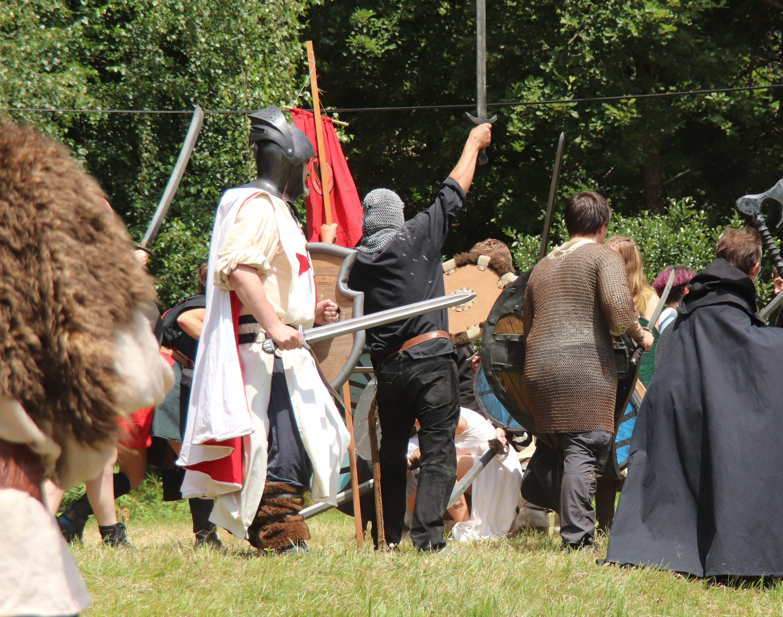 L'image représente une scène de combat. On y voit notamment un chevalier avec un tabard blanc, un homme en noir avec un camail brandissant une épée et un autre homme avec une cote de mail. On peut aussi apercevoir d'autres épées et des boucliers. La scène se passe dans un champs près de la forêt.